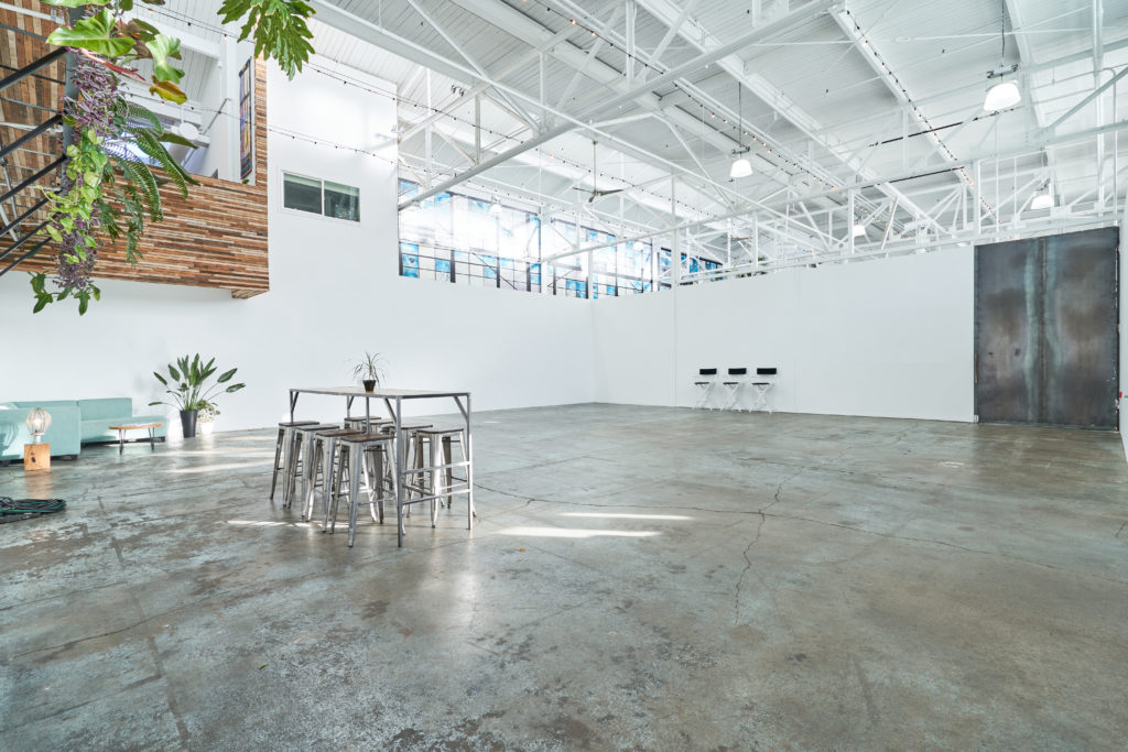Studio 4 is a large, open-concept, multimedia studio space with a private loading dock on the right and three director chairs on the back wall to the right. There are also tall, white walls with exposed white industrial beams on the ceiling. To the left you will see a table with chairs as well as a teal couch with a small coffee table in front.