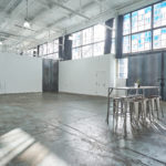 An industrial table with a set of barstool chairs sit in front of a private loading dock on the right of Studio 4. You'll also see a window with some blue panes above the loading dock while 3 black directors chairs sit in the middle of a white wall on the back left of this multimedia studio space.