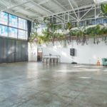 This shot of Studio 4 shows a multimedia studio space with a private loading dock on the left, followed by an open doorway, a small table with chairs, and a large black power box. There is also a teal couch with a table on the right that sits beneath green hanging plants.