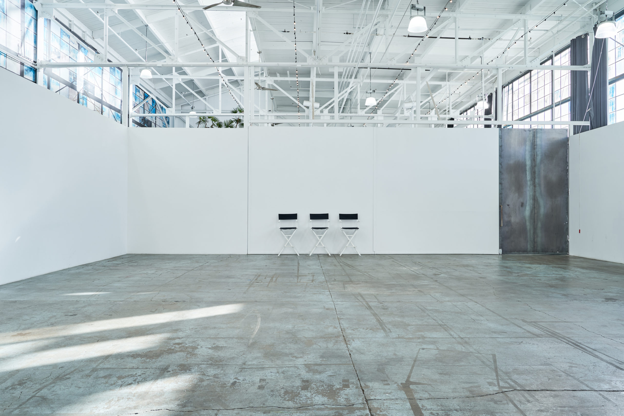 View of Studio 4 with three black director's chairs sitting in the middle of a white wall with industrial white beams on the ceiling and giant windows high up on both sides of this multimedia studio space.