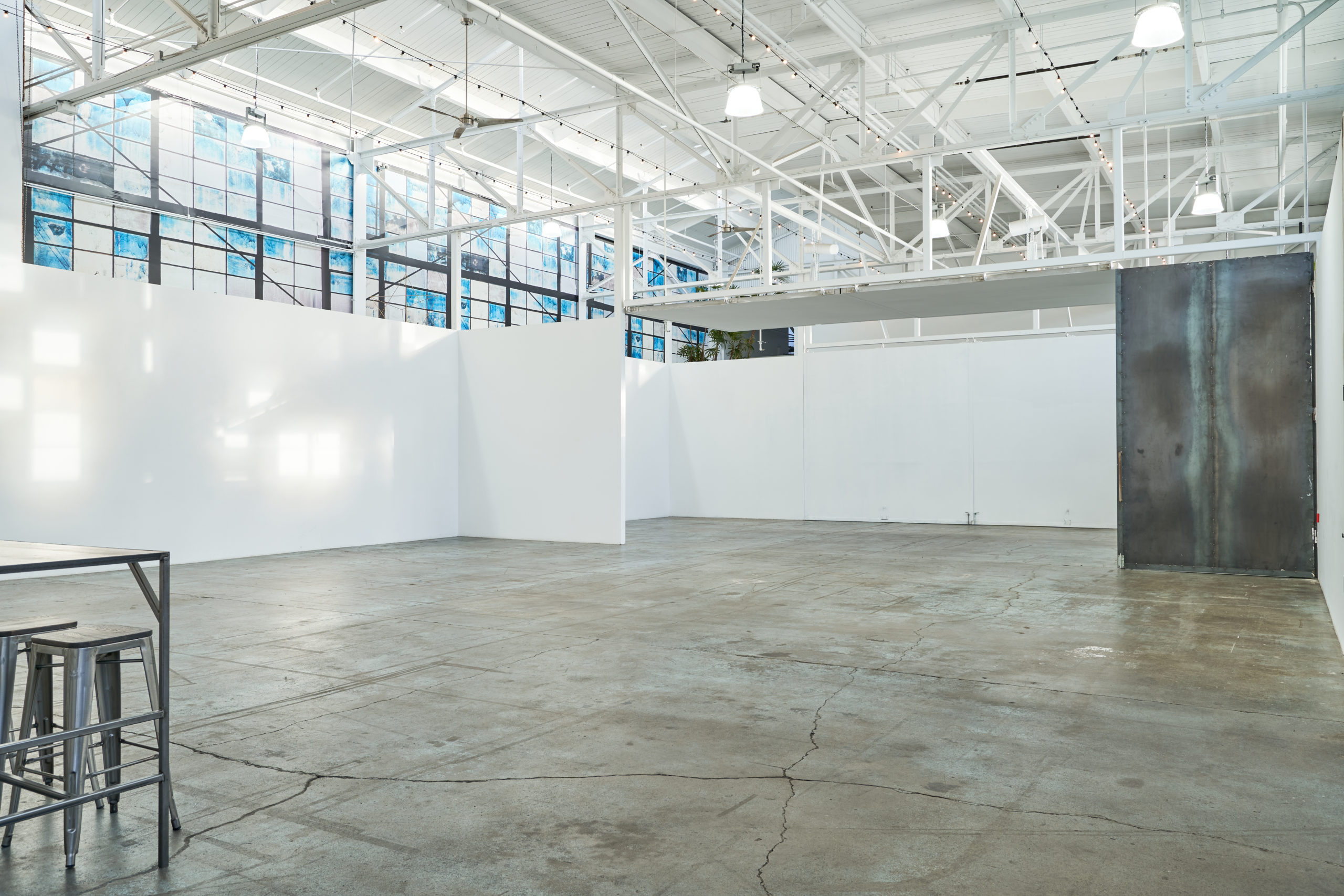 View of the wide open space of Studio 4 with white walls, white industrial beams on the ceiling, and large windows on the top left with blue panes in this multimedia studio space. You can also see a table with chairs in the foreground on the left.