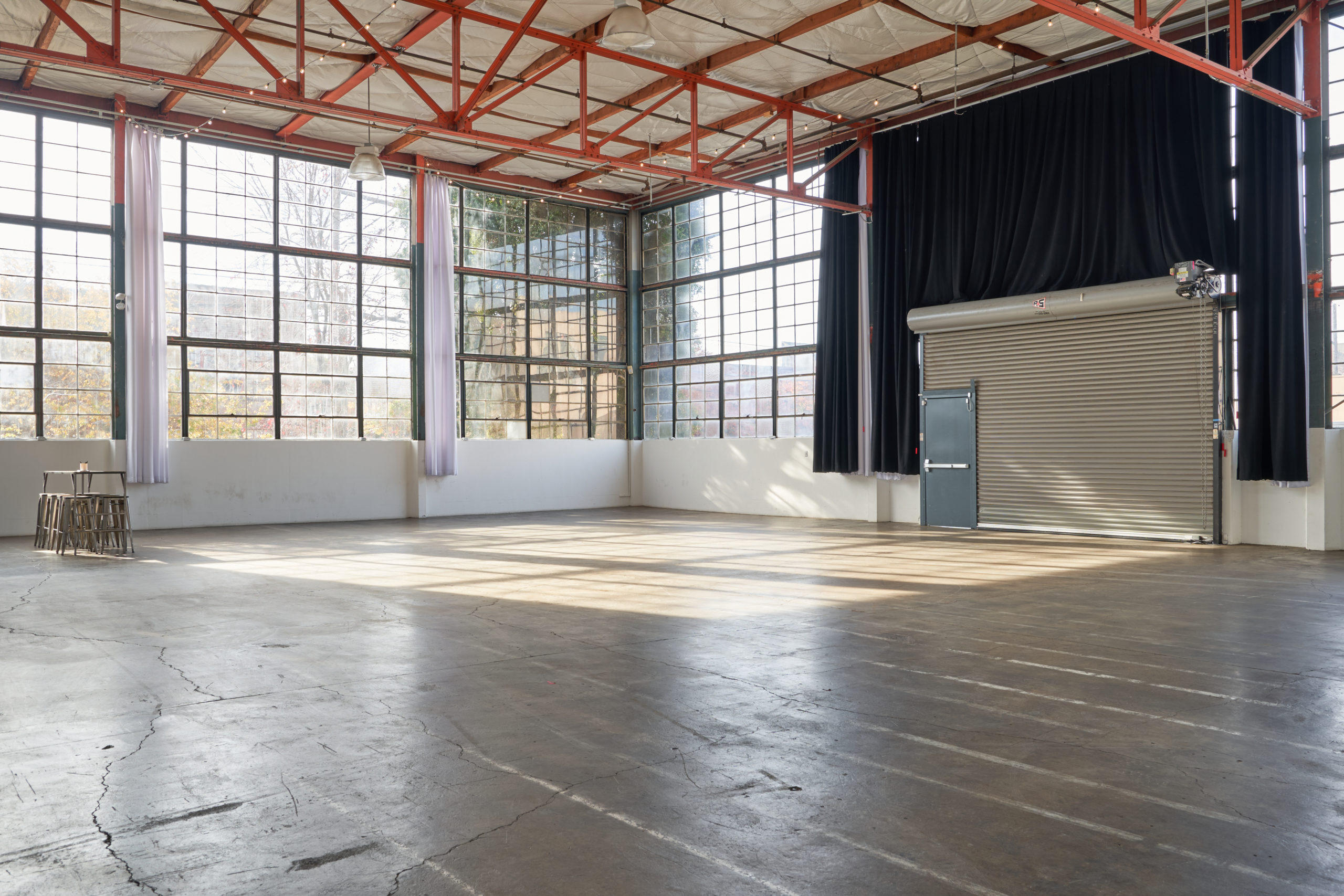 Expansive view of Studio 5 with 3,700 square feet of space. You see a closed door over the private loading dock, a large wall of windows, a small table with chairs, and orange industrial beams on the ceiling of the best multiple set studio rental Berkeley has.