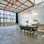 This 2,000 square feet space has lots of light let in from a giant window. It also has a private kitchenette, a ceiling with orange beams, and a large table with seating inside this great creative studio space for rent.