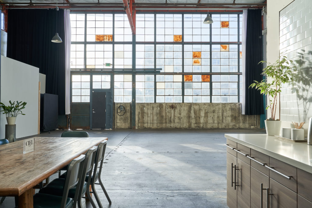 A 2,000-square foot room with a large window with opaque, orange blocks to let in natural light. This creative studio space for rent also has a table with seating and a standing table in the foreground.