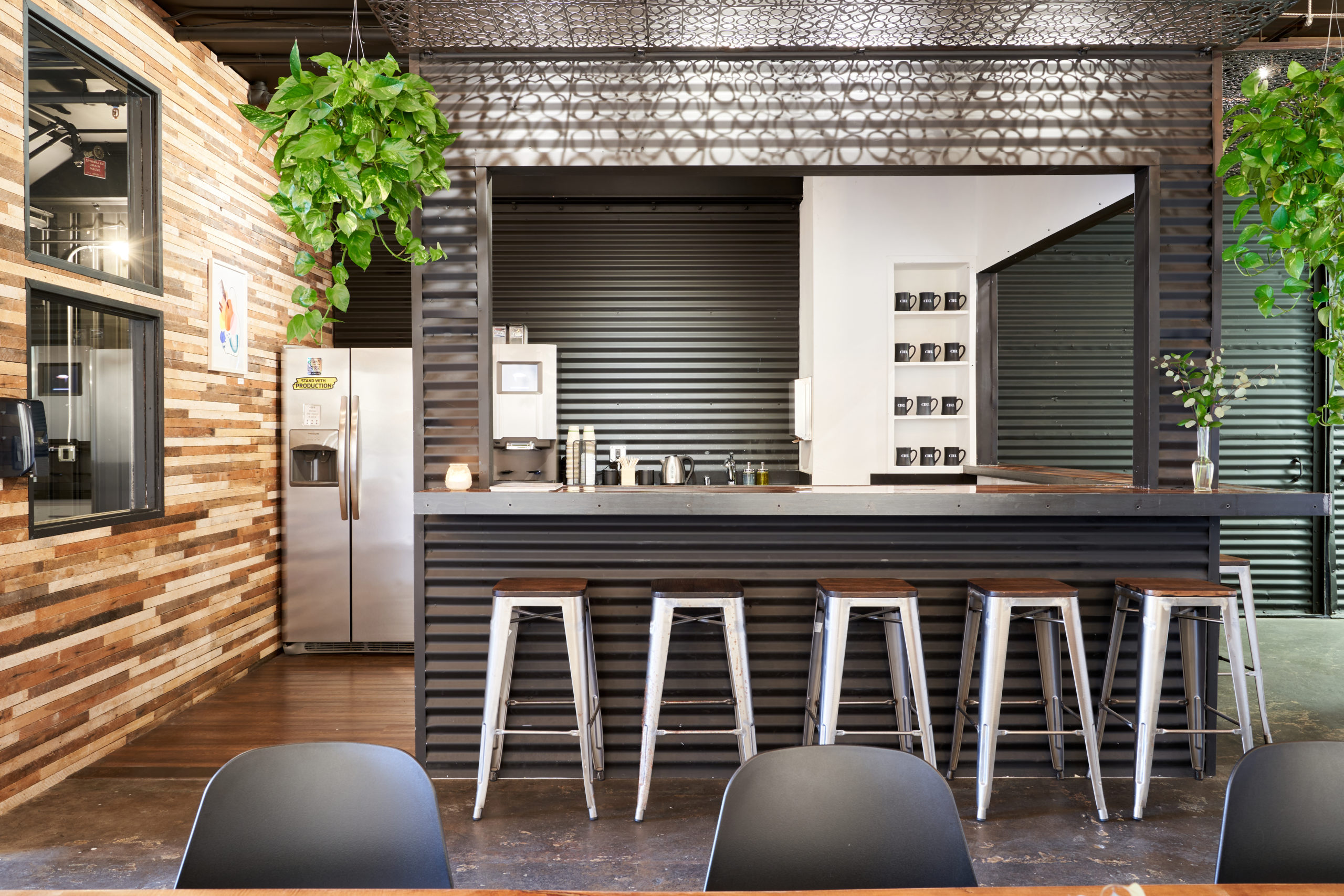 A close up view of the kitchen with a wide open window and five stools that sit at the bar, The kitchen contains a fridage and coffee maker and has black walls with green plants hanging on either side. A perfect addition to the best shared offices Berkeley has.