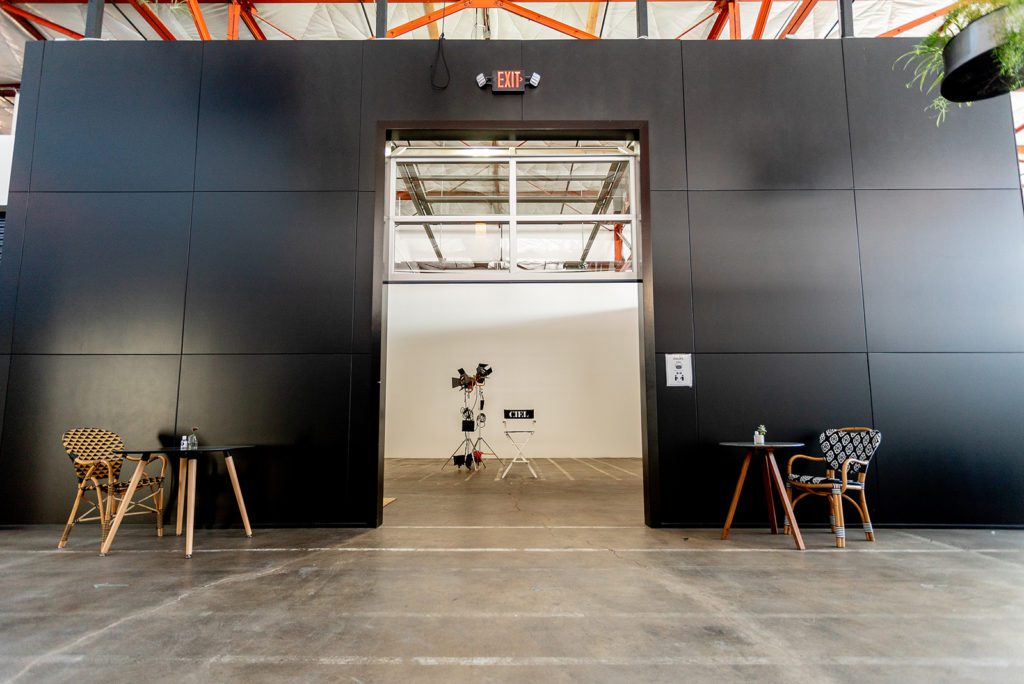 This spacious visual production studio rental features a large entranceway through a black wall that is flanked by a modern table and chair on either side. There is also a large camera inside the bright space of Studio 6.